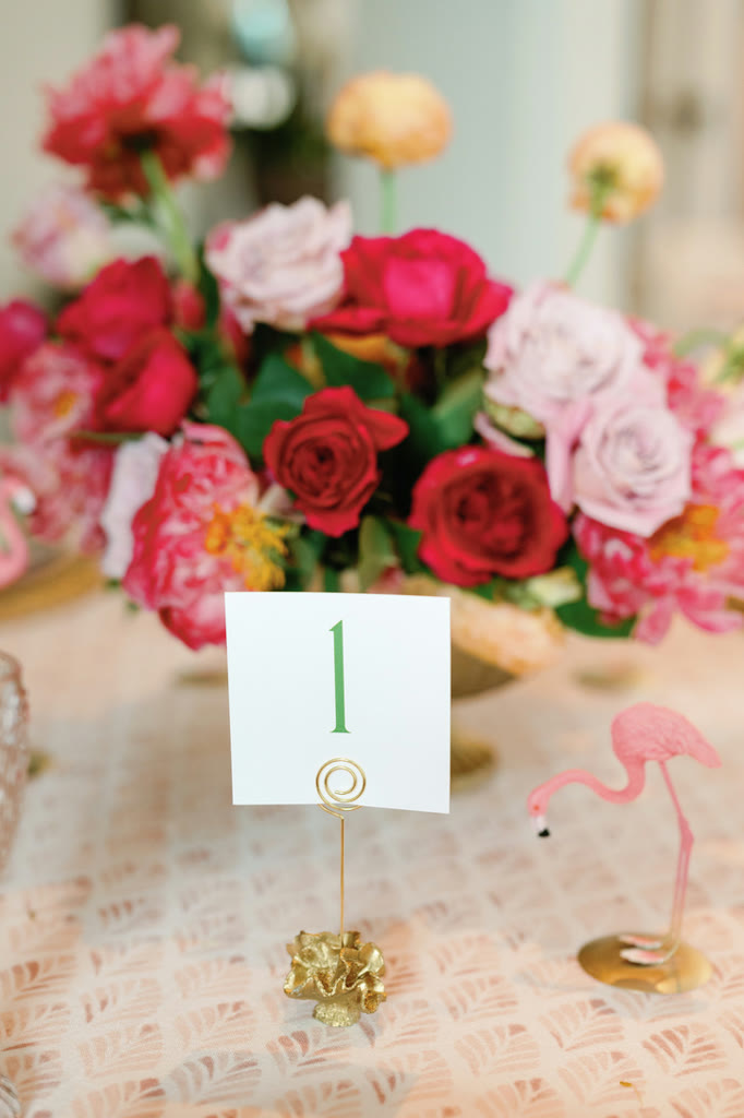 Gold table number card and a pink flamingo place marker in front of a red and pink centerpiece