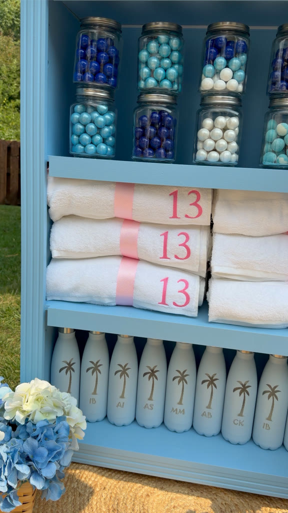 Monogrammed towels, palm-etched water bottles, and candy jars on the shelves of a custom favor cabinet